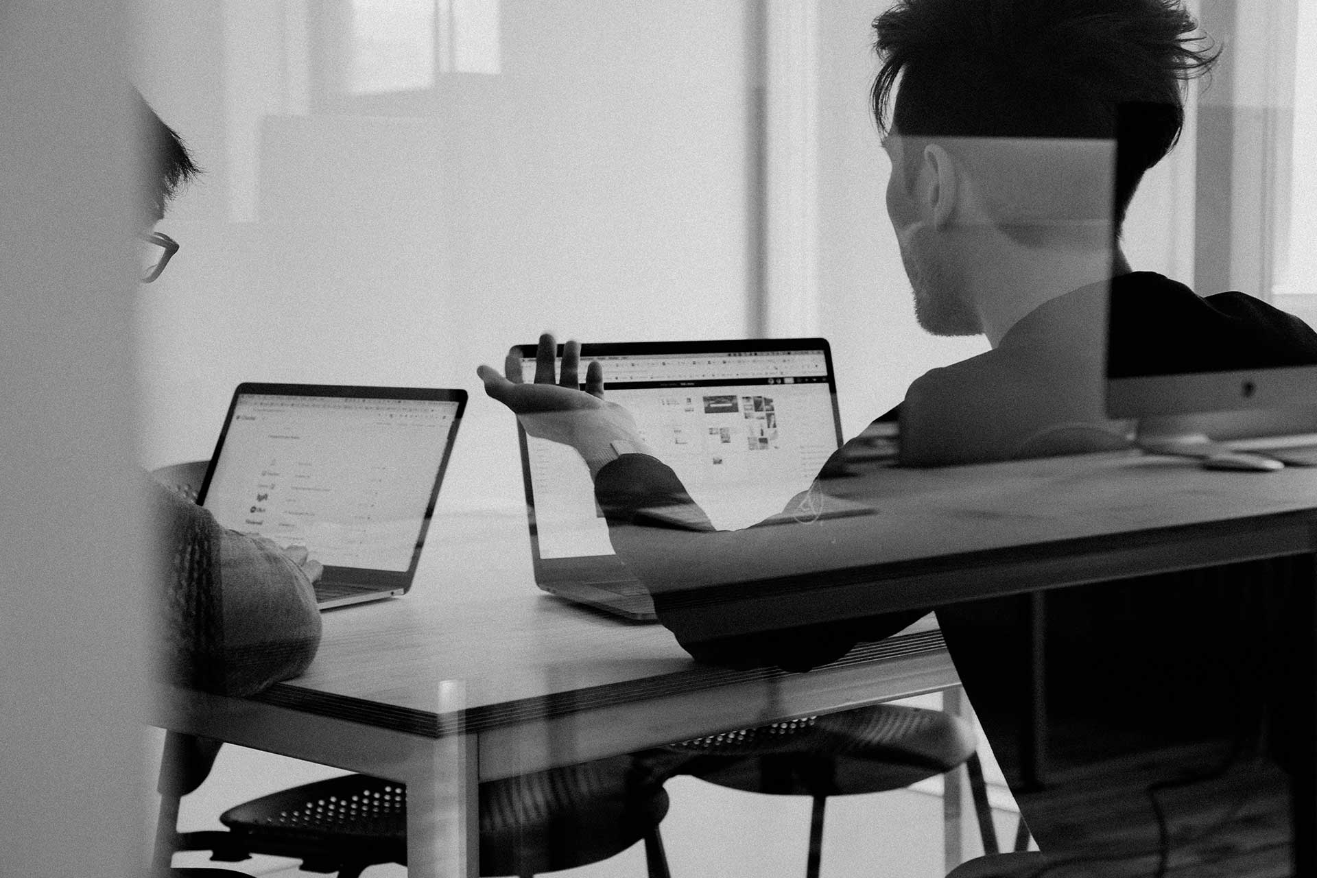 Two people collaborating on laptops at a desk, viewed through an office partition.